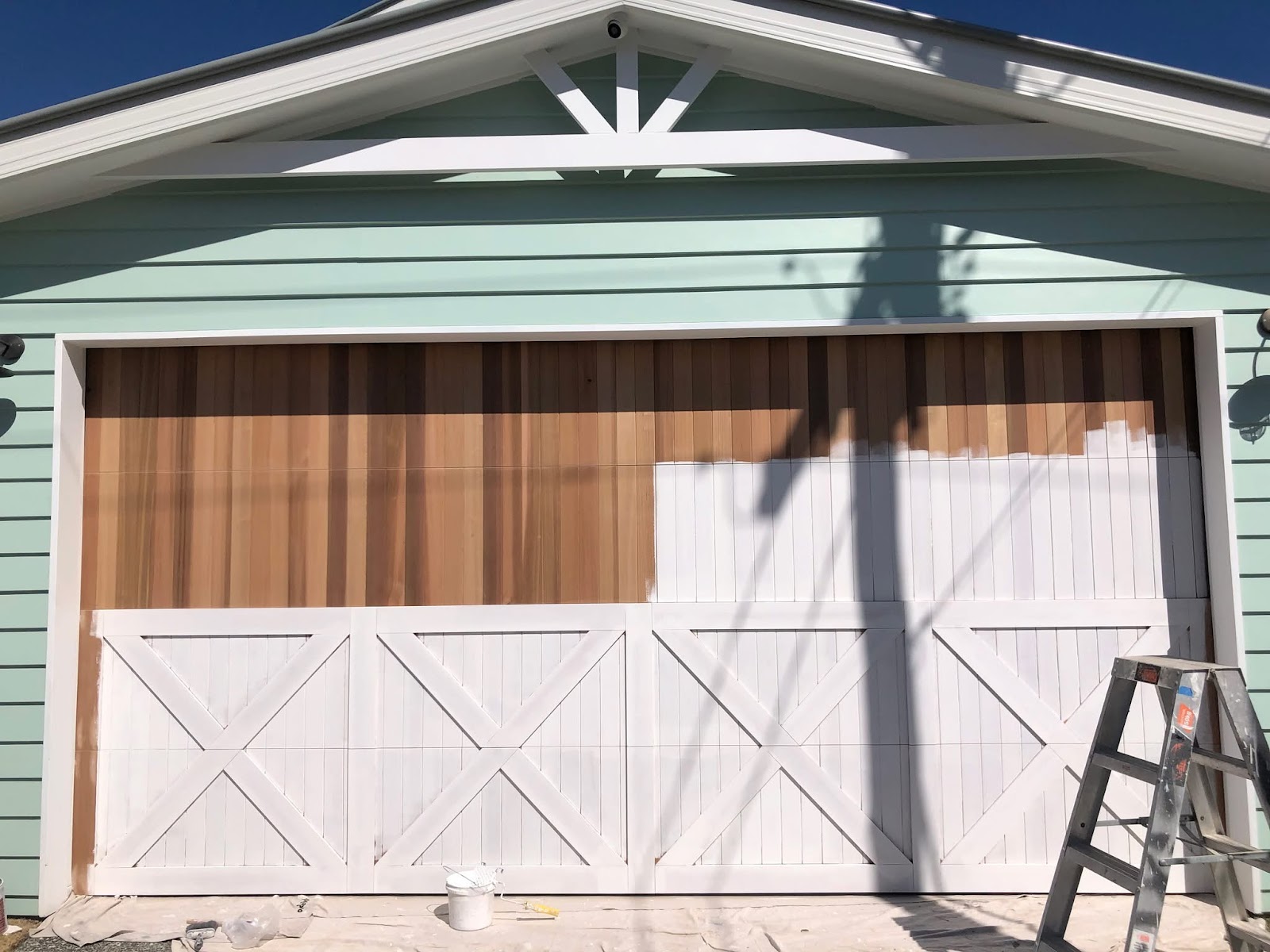 Sage green weatherboard house with a timber garage door being painted white, ladder beside it